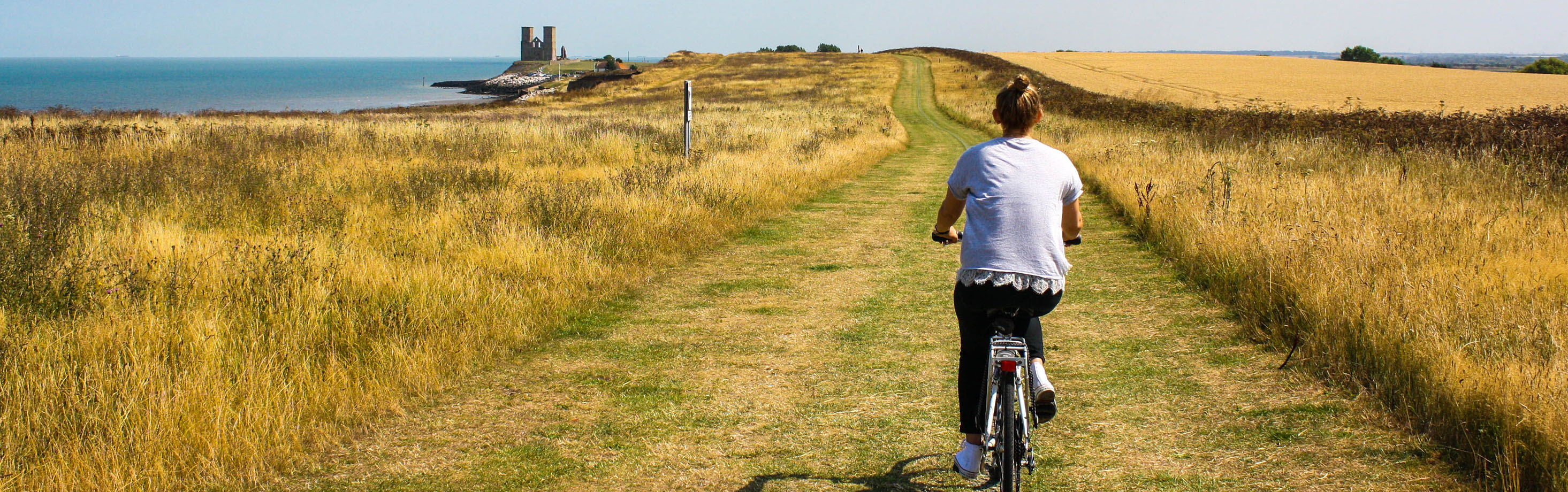 A person on a bicycle rides along a narrow dirt path through a golden field of tall grass, with the ocean and a distant building visible on the left.