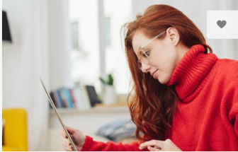 A woman with long red hair, wearing a red turtleneck, smiles while looking at a tablet in her hands.