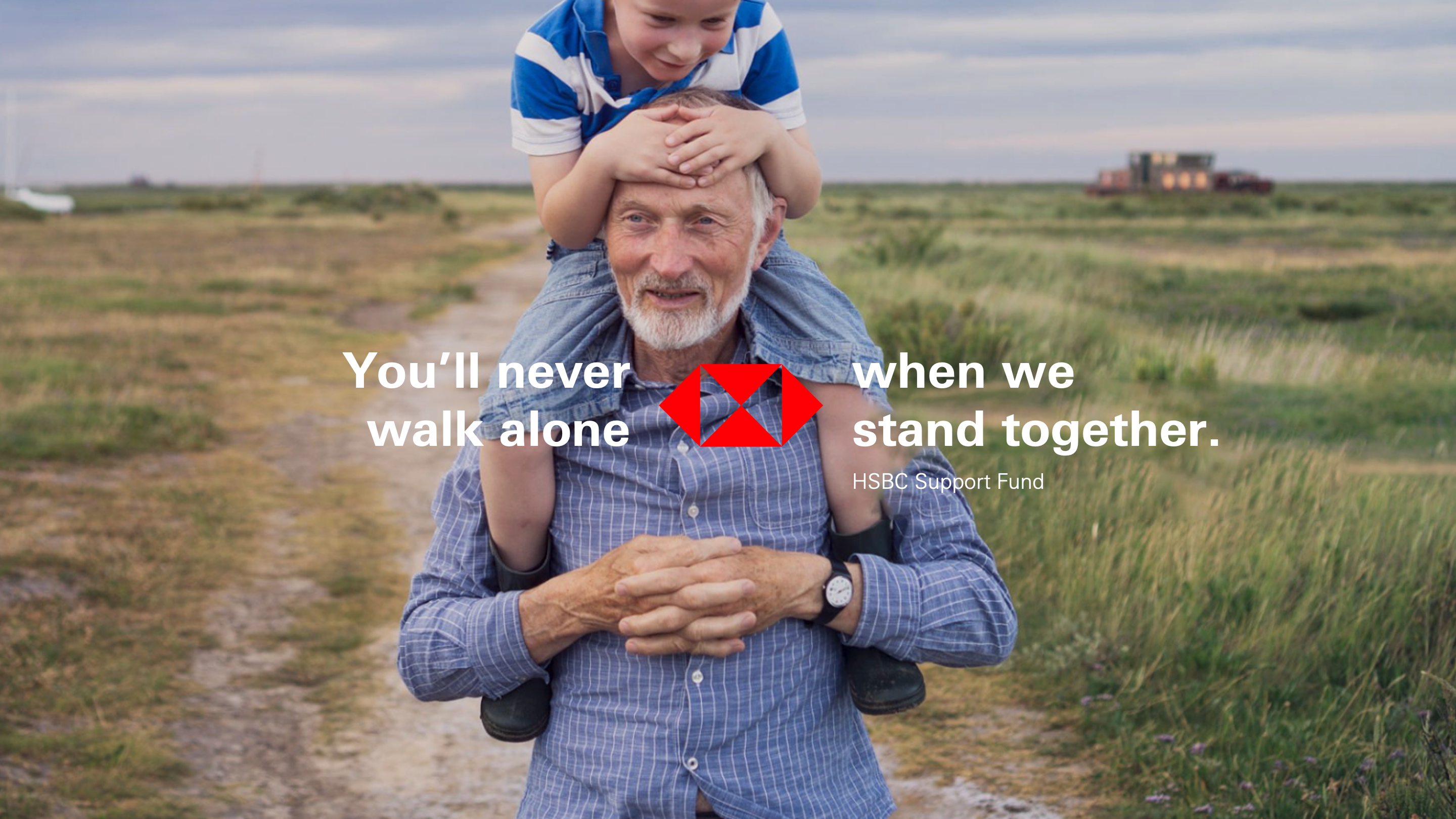 A young boy sits on the shoulders of an older man, both smiling, walking on a dirt path in a rural setting with text overlay "You'll never walk alone when we stand together. HSBC Support Fund".