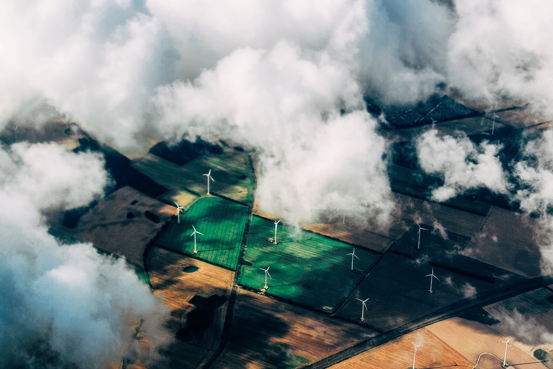 An aerial view of green and brown fields dotted with wind turbines, partially obscured by fluffy white clouds.