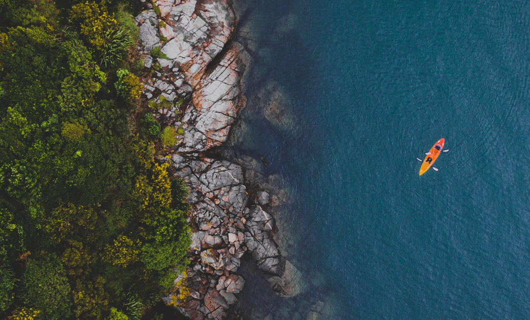 An aerial view of a rugged coastline with dense green forest meeting dark blue ocean water, and a small orange kayak visible on the water.