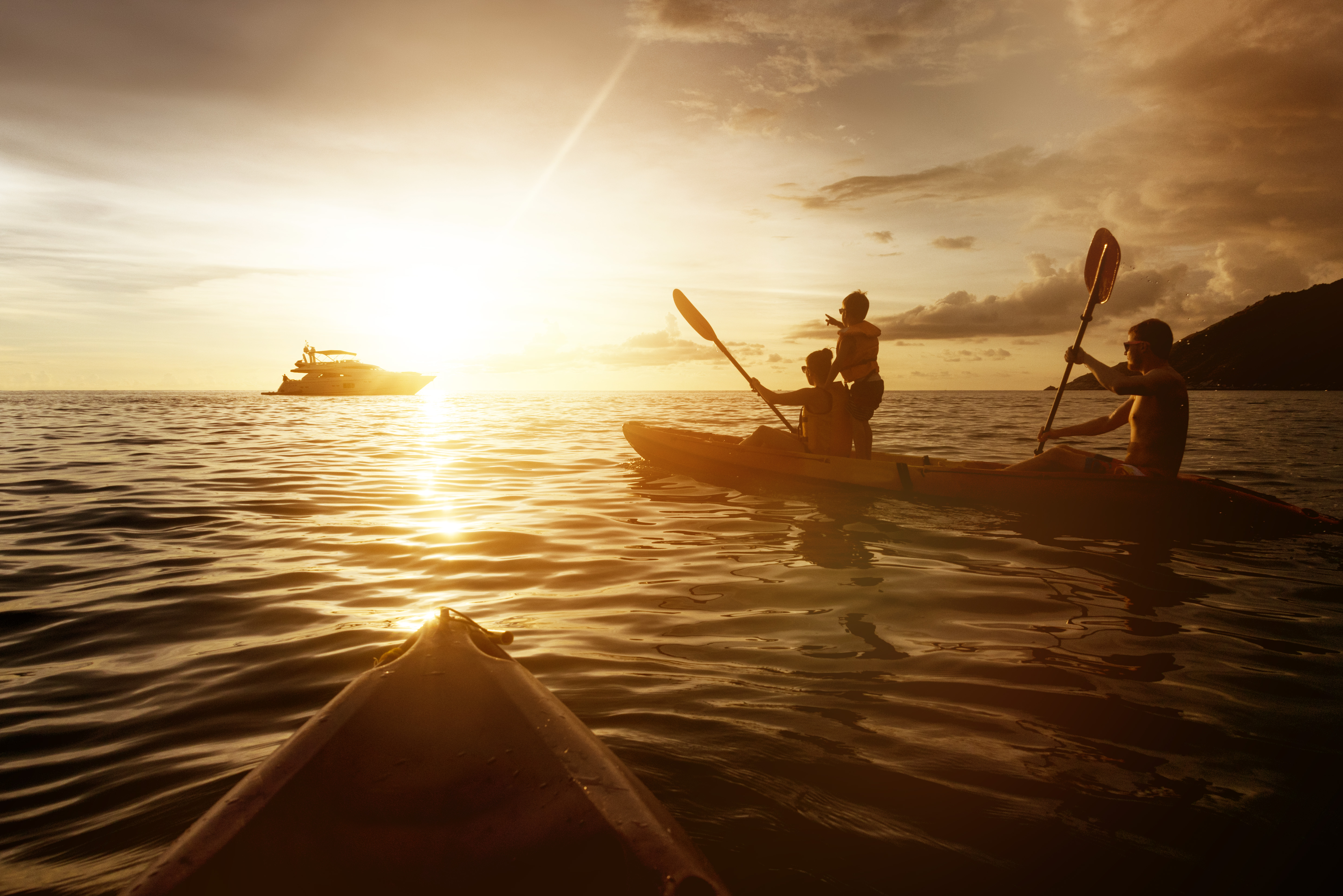 Two people in a kayak on calm water at sunset, with a motor yacht in the distance and a third person standing in the kayak pointing towards the horizon.