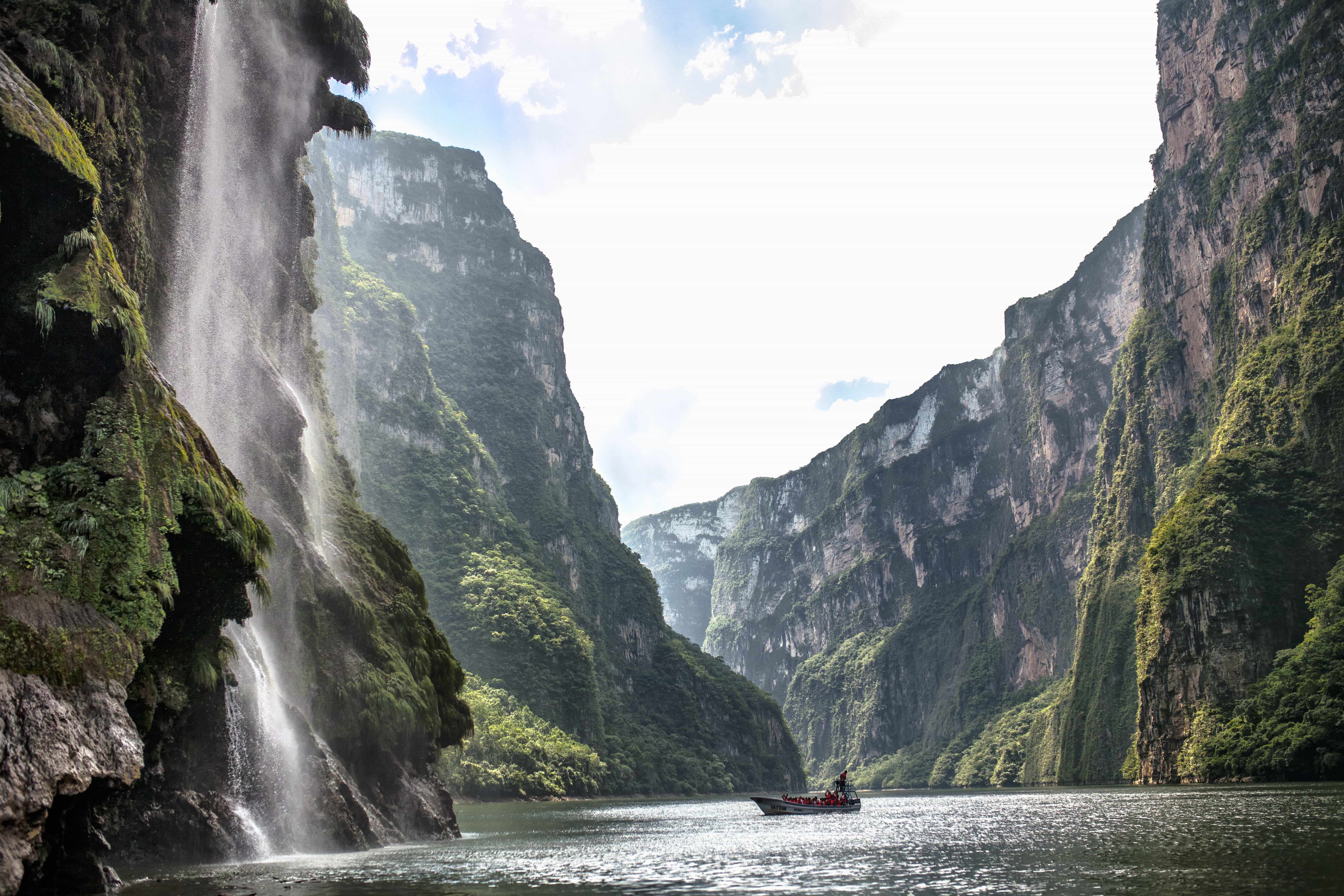 A wide river flows through a deep canyon with tall, green-covered cliffs and a large waterfall cascading down one side, under a bright sky. A small boat is on the water.