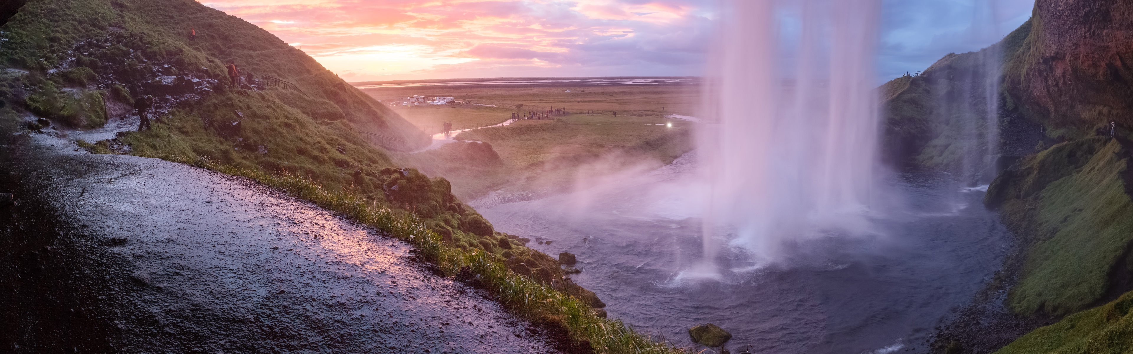 A wide waterfall cascades over a cliff into a river, with a pink and orange sunset sky in the background.