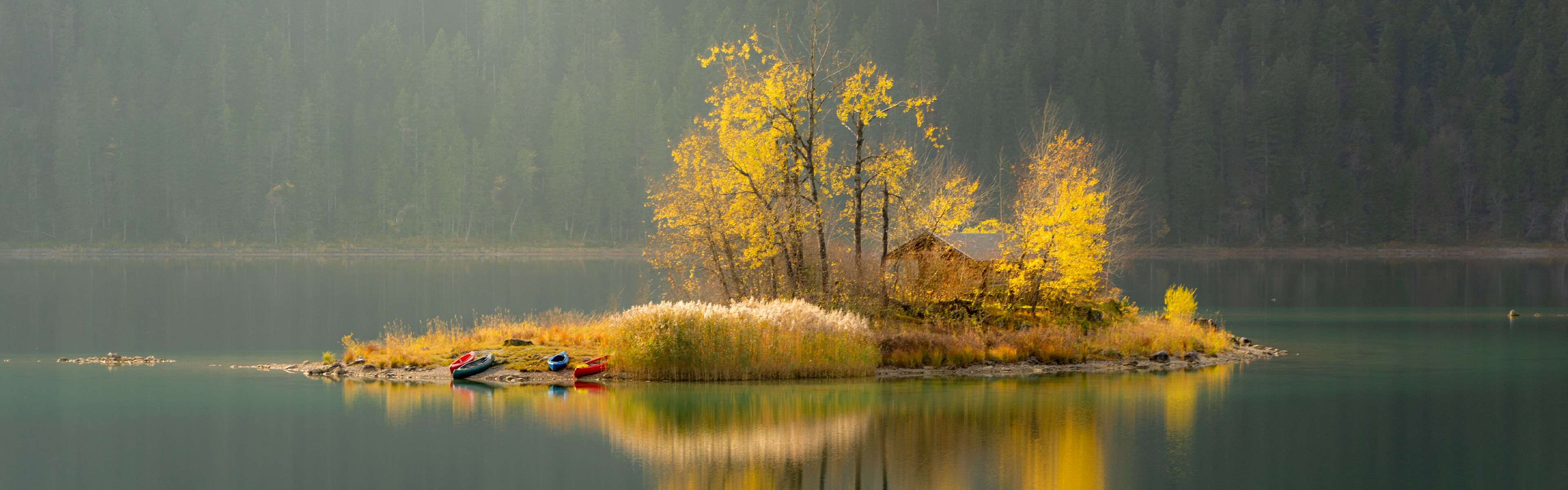 A small, tree-covered island with a few colorful boats docked, sits in a calm lake surrounded by autumn trees under a hazy sky.