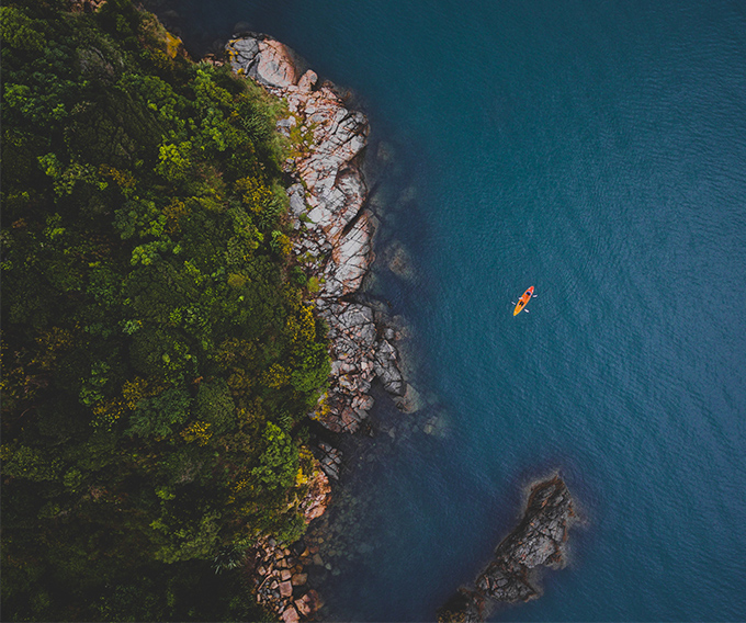 An aerial view of a rugged coastline with dense green forest meeting dark blue ocean water, and a small orange kayak visible on the water.